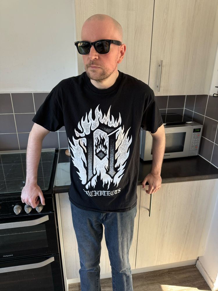 A man standing in a modern kitchen. He is bald, has a short beard, and is wearing dark sunglasses. His outfit consists of a black T-shirt with a bold white graphic design featuring flames and the word "ARCHITECTS" at the bottom, along with blue jeans. He has one hand resting on the handle of an oven and the other on a kitchen drawer handle. There's a microwave on the countertop behind him, and the room has light wood cabinets and dark countertop surfaces. The lighting in the room is bright, likely from sunlight.