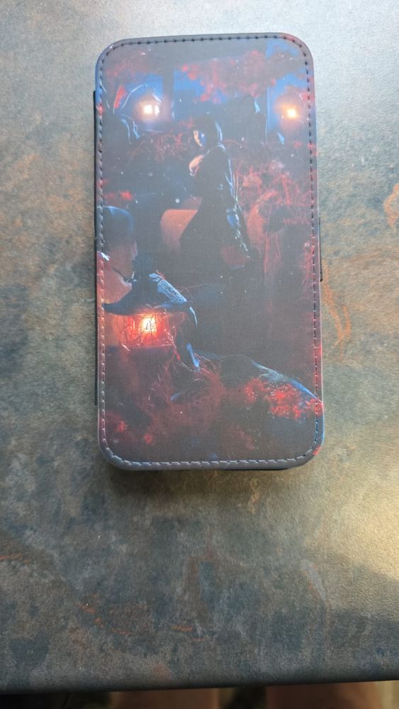 The design on the phone case is a detailed, moody artistic scene dominated by deep blues, reddish hues, and glowing orange lights. Here are the specifics:

The image appears to take place in a forest or a wooded setting at night. The background features blurry red foliage, possibly autumn leaves, and two distinct glowing lanterns or lamps high up, casting a warm, golden light that contrasts against the dark blue and black surroundings.

In the middle of the case, there is a prominent figure—likely a woman—wearing a long cloak or coat. She’s standing beside or above another figure who seems crouched or kneeling, possibly holding something. The main character looks over her shoulder, and her face is softly lit by the surrounding glow, giving her a mysterious but calm expression.

Below, there’s another light source, this time from a lantern near the ground, partially obscured by twisted roots or branches outlined in red light. The lower half of the case is darker but features more reddish-orange highlights along roots or tendrils, creating almost a web-like or magical effect around the lantern.

The overall illustration has a fantasy or supernatural vibe, reminiscent of scenes from dark fairy tales or graphic novels. The sewing along the case's border frames the image neatly. The colors, lighting, and elements all contribute to a mysterious, enchanting nighttime atmosphere.