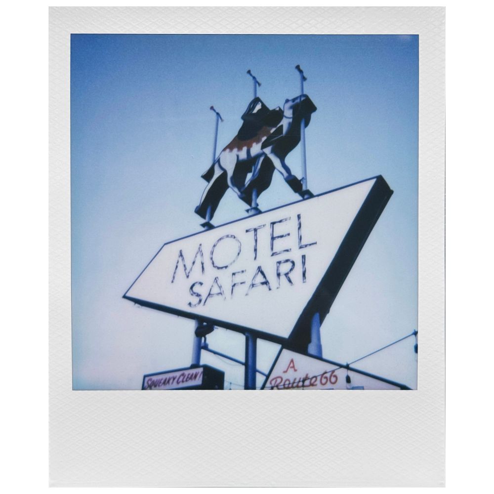Polaroid photograph of the sign for the vintage Motel Safari with a blue sky in the background. It features a person riding a camel placed atop an angular piece with the name of the motel on it. 