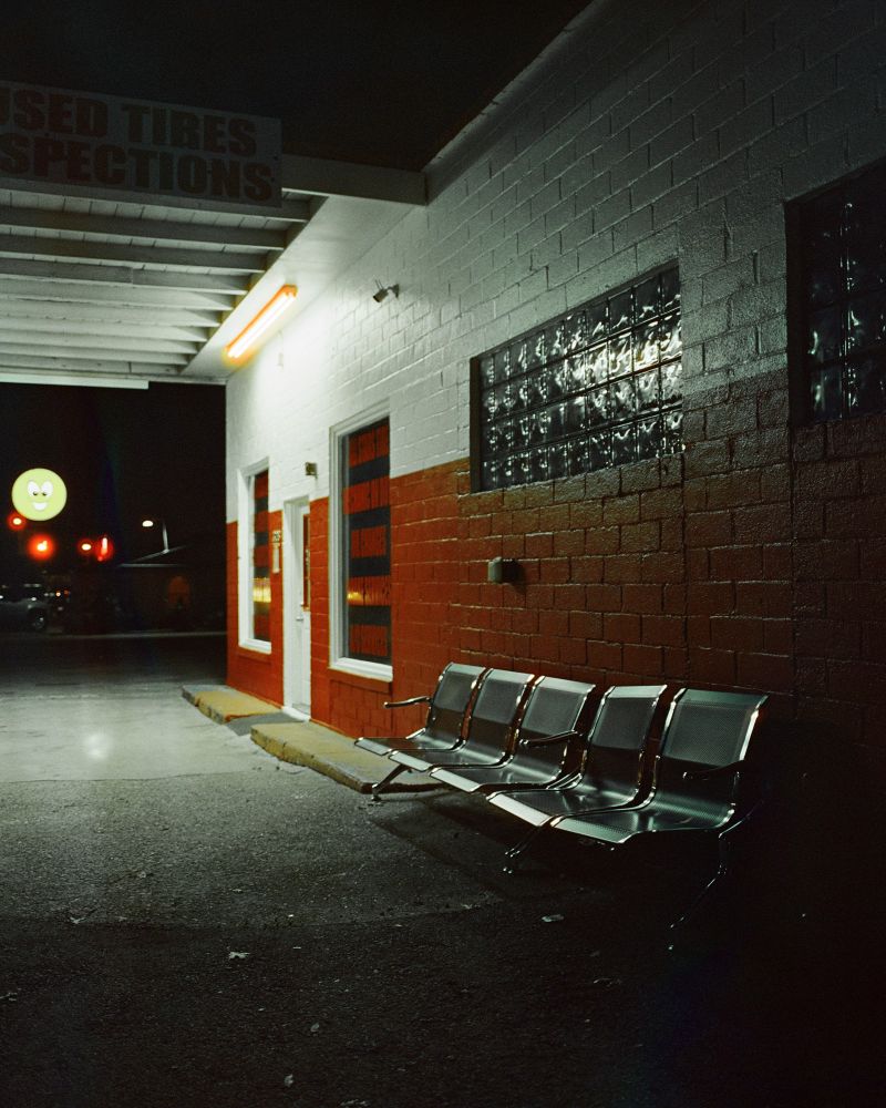 Photograph of a night scene outside of a tire repair shop. The front of the brick building is painted red and a white, and a single long fluorescent light illuminates a row of chairs near the door which serve as a waiting area. 