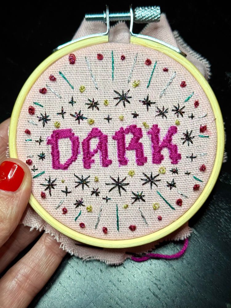A hand with a red-painted fingernail holds a small, round embroidered frame on which the word DARK is embroidered in medieval pink lettering. Surrounded by numerous stars, dots and rays of light made from glittering yarn in black, pink, silver, gold and turquoise.