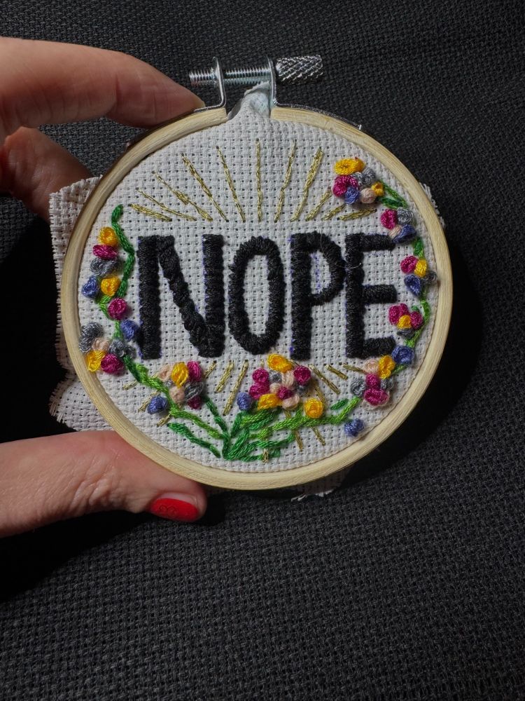 A hand holding a small round embroidery hoop with the lettering NOPE on it. Golden rays and colourful flower tendrils are embroidered around it.