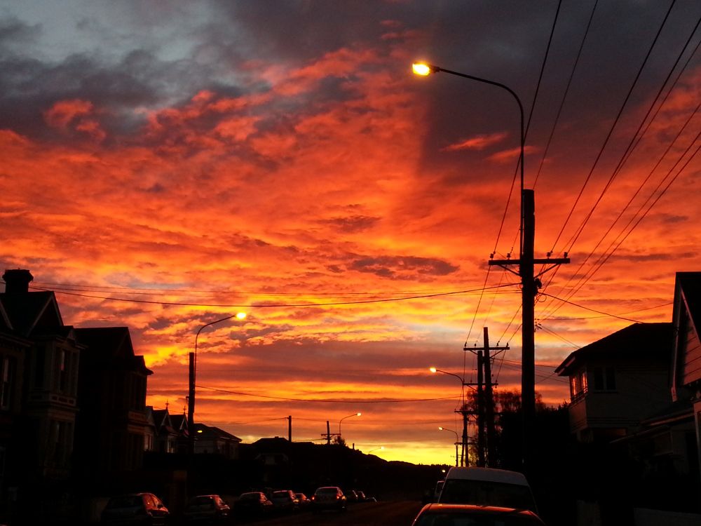 Sunrise over a street with cars on each side. The frosty morning can be seen on how the light reflects on the car roof tops. Sky is covered in clouds that are a strong red and gold colour