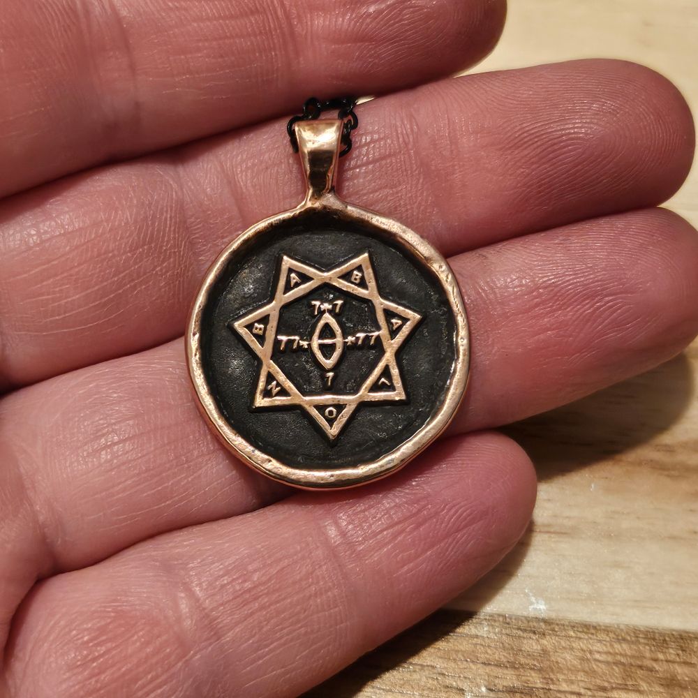 A white person's light pink fingers hold a copper pendant with a triangular bail on a black sterling steel chain. The pendant is round and a little larger than a quarter with a bright copper border and seal of Babalon both rising up from the recesses of the pendant. The recesses have a dark brown-black patina from liver of sulfur.