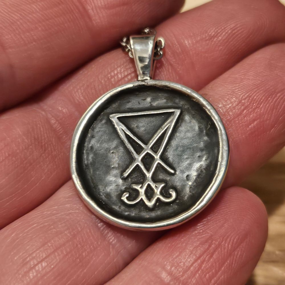 A white person's light pink fingers gold a fine silver pendant with a triangular bail on a sterling steel chain. The pendant is round and a little larger than a quarter with a bright silver border and sigil of Lucifer both rising up from the recesses of the pendant. The recesses have a dark gray patina from liver of sulfur.