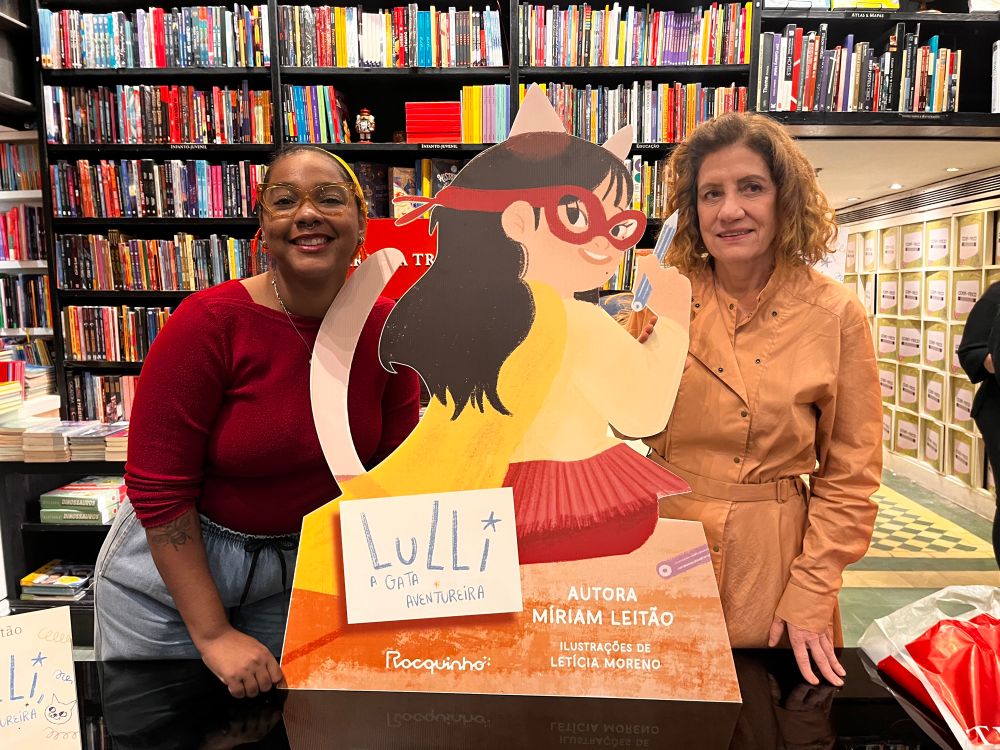 Imagens de Letícia Moreno e Miriam Leitão, junto de um totem com a personagem do livro, Lulli