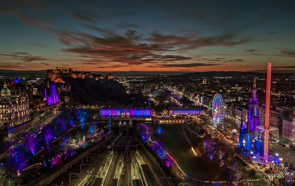 Aerial night shot of the sunset over Edinburgh in distance with lights from Christmas markets LNER Big Wheel and star flyer in foreground 