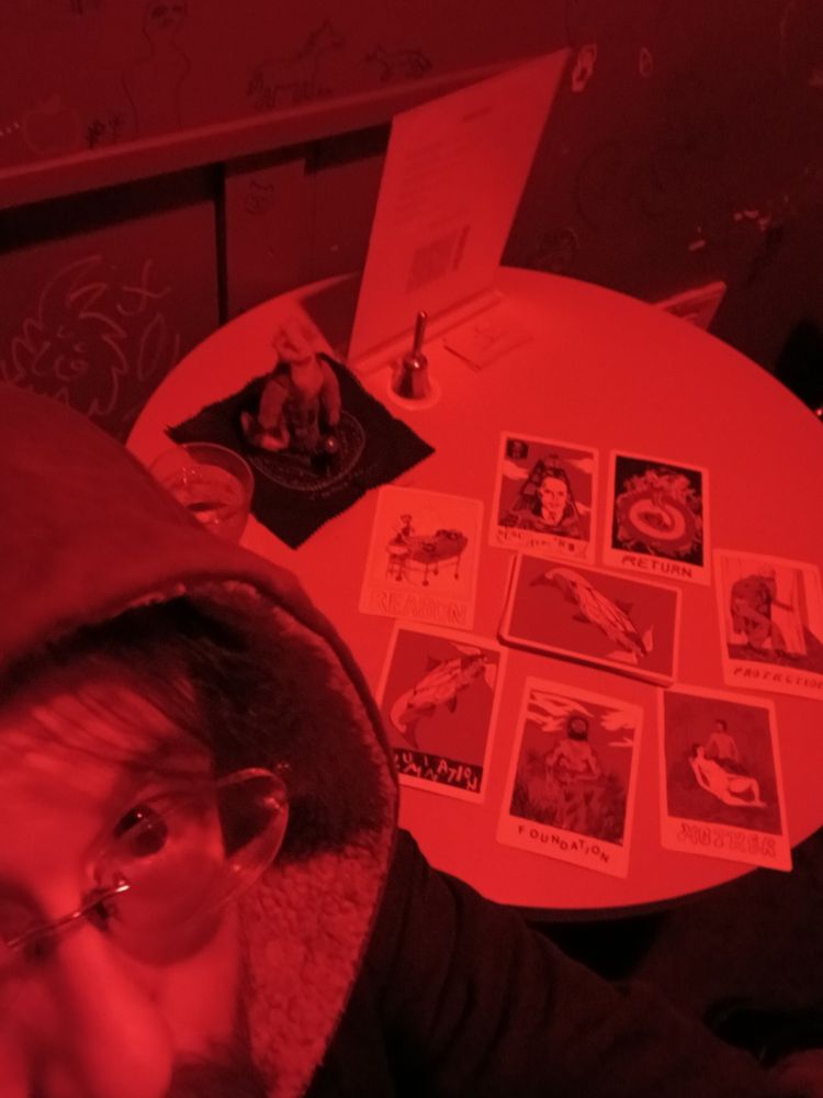 A cartomantic spread on a table lit by a red light. A hooded Connor kizer peaks in the lower left corner.
