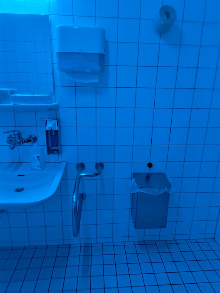 Bild des Waschbeckens einer barrierefreien Krankenhaus-Toilette. das Ersatztoilettenpapier ist ist knapp 2m Höhe an der Wand hängend befestigt. Der Papierhandtuchspender in ca. 1,7m Höhe. Der Raum ist in Blaulicht beleuchtet.