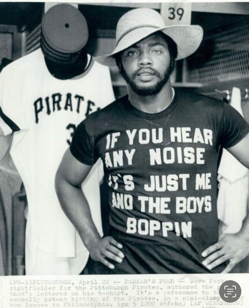 Dave Parker standing with his hands on his hips in front of an open locker with a “39” placard and a Pirates uniform and hat on a hook. He is wearing a sun hat and a black t-shirt with the words IF YOU HEAR ANY NOISE IT’S JUST ME AND THE BOYS BOPPIN.