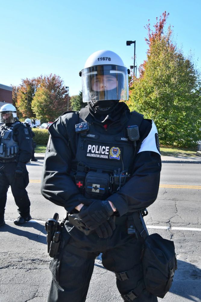 Antiémeute de face avec casque et gilet pare balle arbirant le symbole du thin blue line qui est un drapeau canadien traversé par une ligne bleue. Brassard blanc.