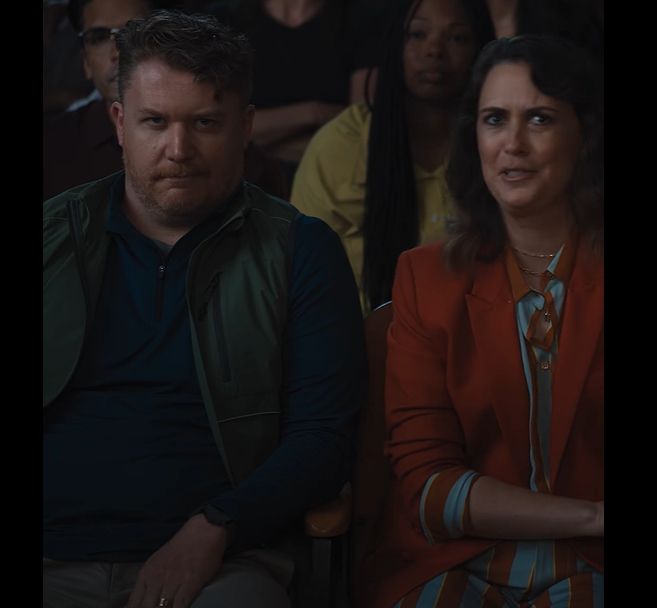 A White mom (the mom of the girl child in the previous photo) sitting next to a White dad (the dad of the boy child in the previous photo), in the audience of the chess tournament