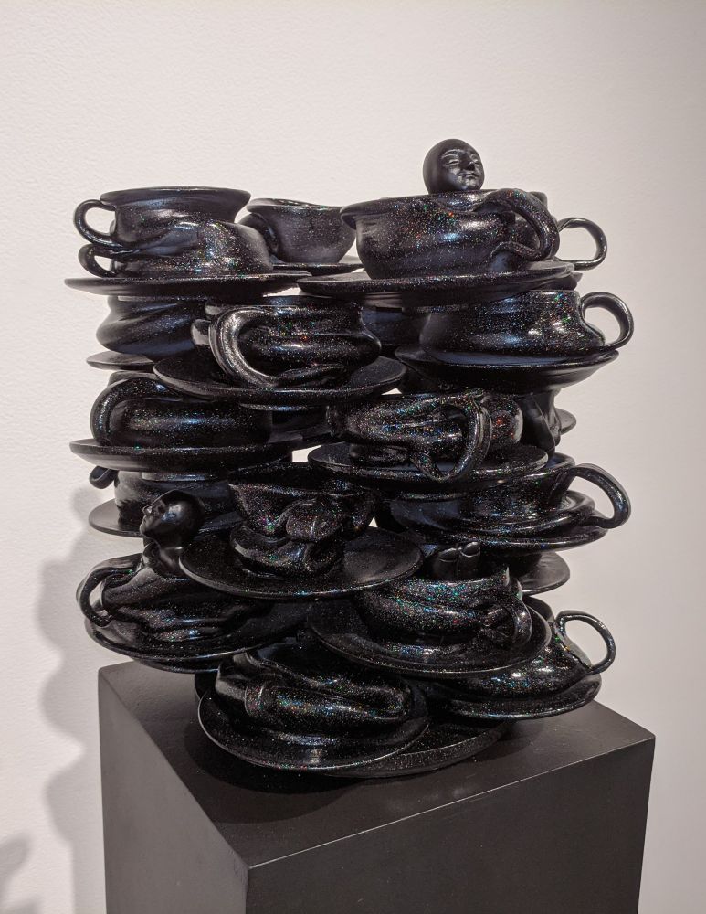 A black glittery sculpture comprised of 24 squashed coffee cups and saucers stacked in groups of 4. There is a head peeking over the rim of one of the top cups.