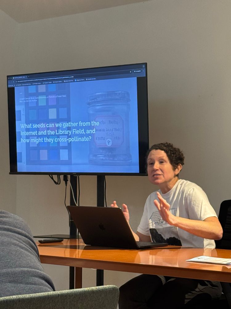 shannon mattern in a white tshirt sitting at a table in front of a laptop presenting to a group in front of a presentation slide that says “what seeds can we gather from the internet and the library field wnd how might they cross pollinate?” The text is on top of a semi opaque background of a color swatch and a small glass jar. 