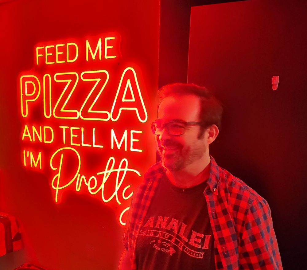 Extremely handsome man standing in front of a red neon sign that reads "Feed me pizza and tell me I'm pretty" at a pizza restaurant 