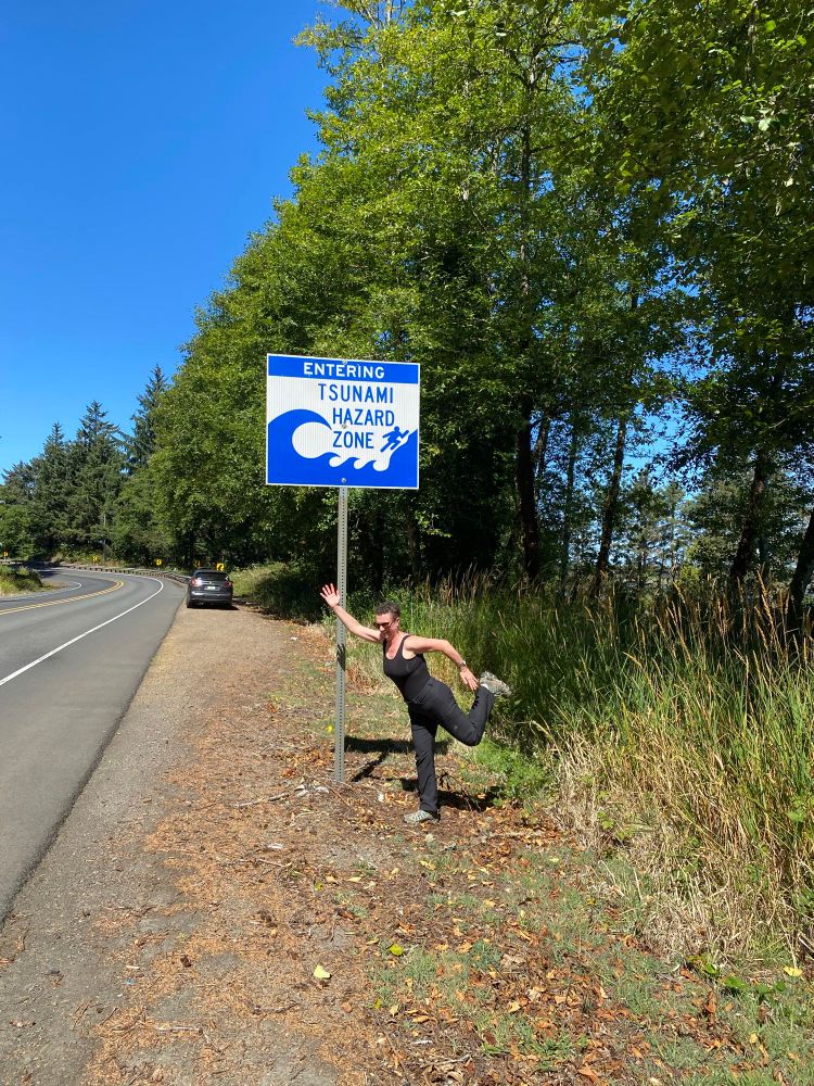 Me, a person with short curly hair wearing dark grey hiking pants and a black tank top, in front of an "Entering Tsunami Hazard Zone" road sign mimicking the pose of the stick figure attempting to escape catastrophe. 