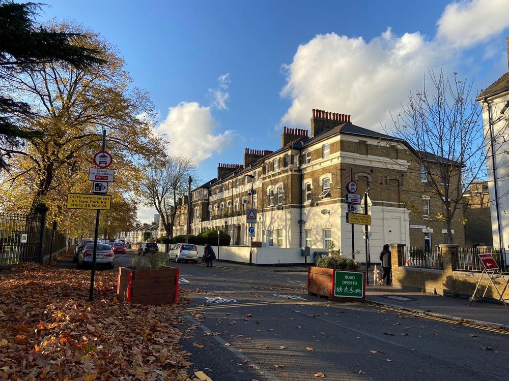 A residential street which has been narrowed with wooden planter boxes at a junction, and traffic signs saying “no motor vehicles except permit holders WHP01, CCTV enforcement on Ham Park Road starts 25 Nov 24.” The street is nice and peaceful with a pedestrian crossing on the corner comfortably. The late afternoon sun casts a golden light on fallen leaves. There is no through traffic to be seen.