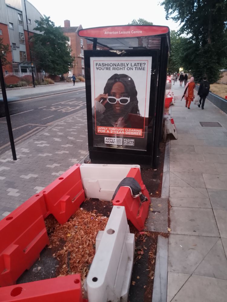 An unfinished bus stop bypass (floating bus stop) at a cycle track, with the bus shelter for the Atherton Leisure Centre still inside the one-way cycle track, waiting to be moved onto the island which is otherwise finished. An advert from the bus shelter stares back at us: "Fashionably late? You're right on time. Apply through clearing for a 2-year degree."