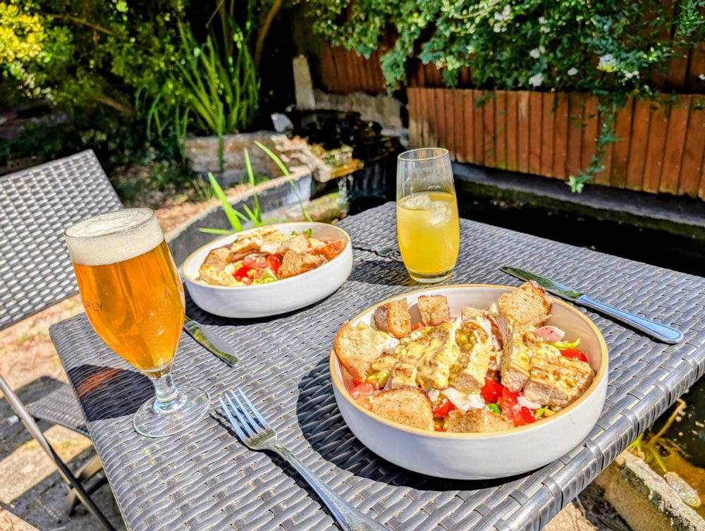 Two bowls of Caesar salad, a glass of beer and a glass of Malibu and pineapple on a wicker table next to a pond. The sun is shining and the vegetables are colourful! 