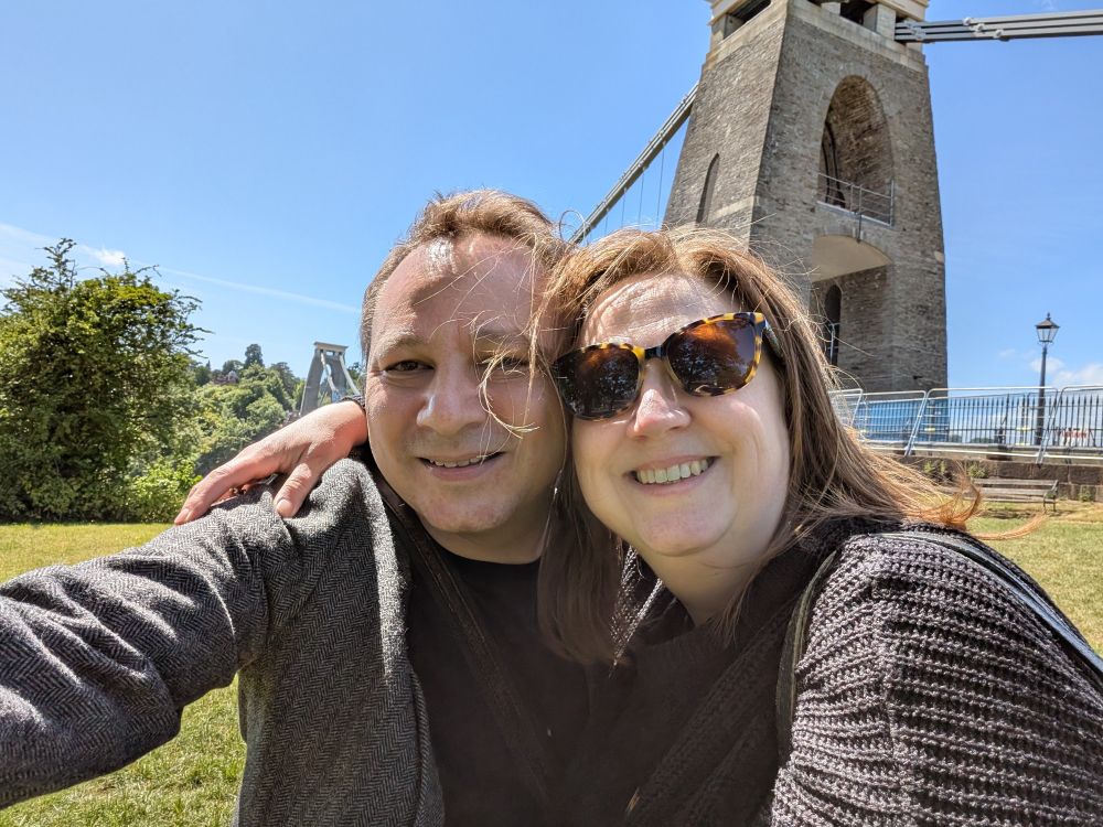 Matt and Lucy with Clifton Suspension Bridge behind. It is a sunny day with blue sky. 