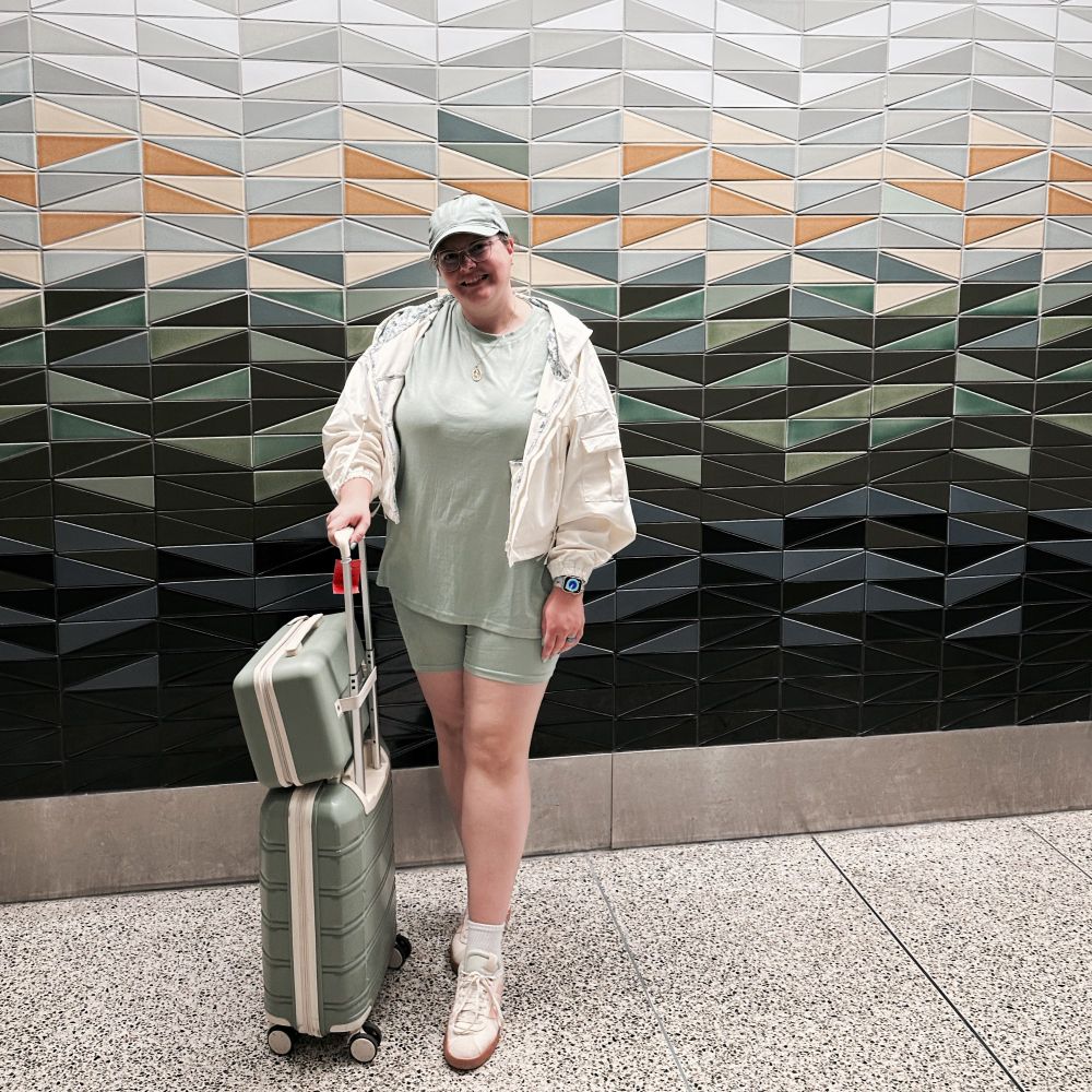 Woman in a matcha green tee and short set, matcha green ball cap, matcha green luggage, with a white windbreaker standing in front of a geometric tiled wall with shades of green, gray, and white with pops of a yellow-orange. 