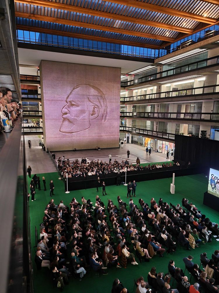 A seated audience in the atrium of the Bell Works aka Lumon building with a massive wall with a relief of Kier projected on to it. 