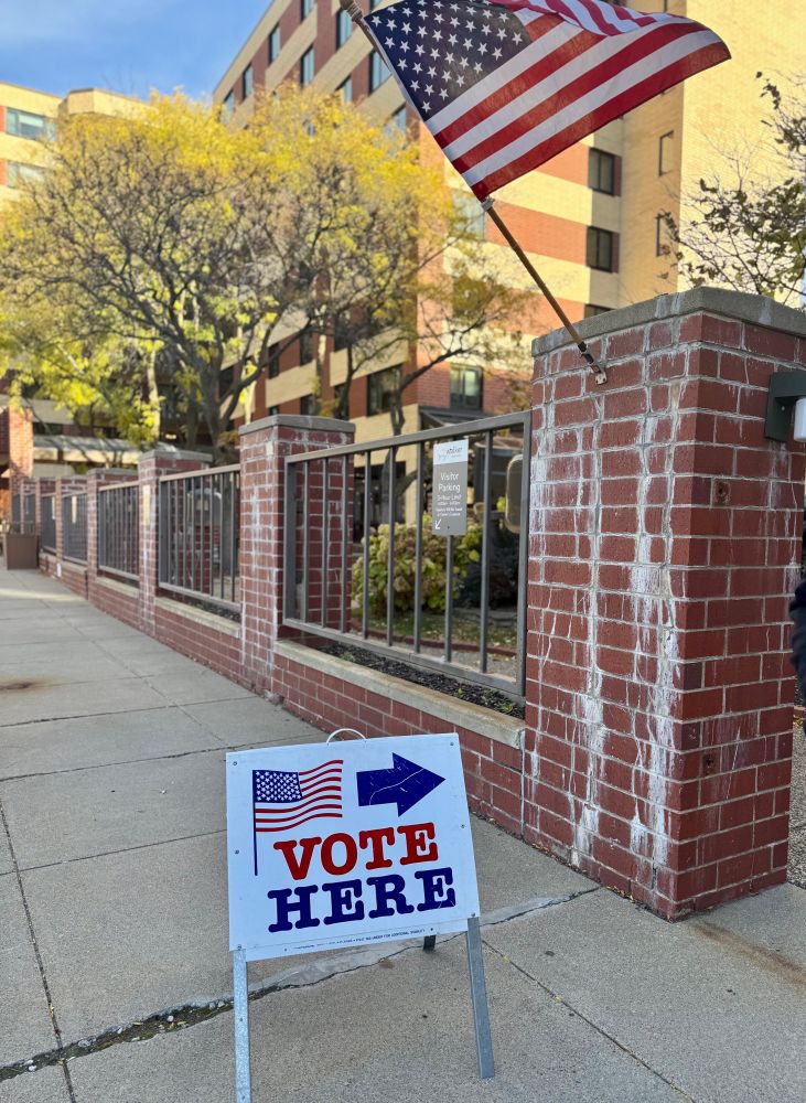 A sidewalk with a Vote Here sign and American flag affixed to a brick fence.