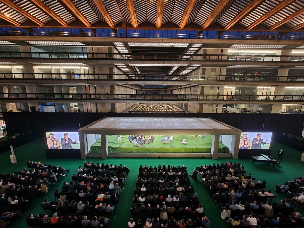 A seated audience in the atrium of the Bell Works aka Lumon building looking at a room with the creators and actors of Severance in it. 