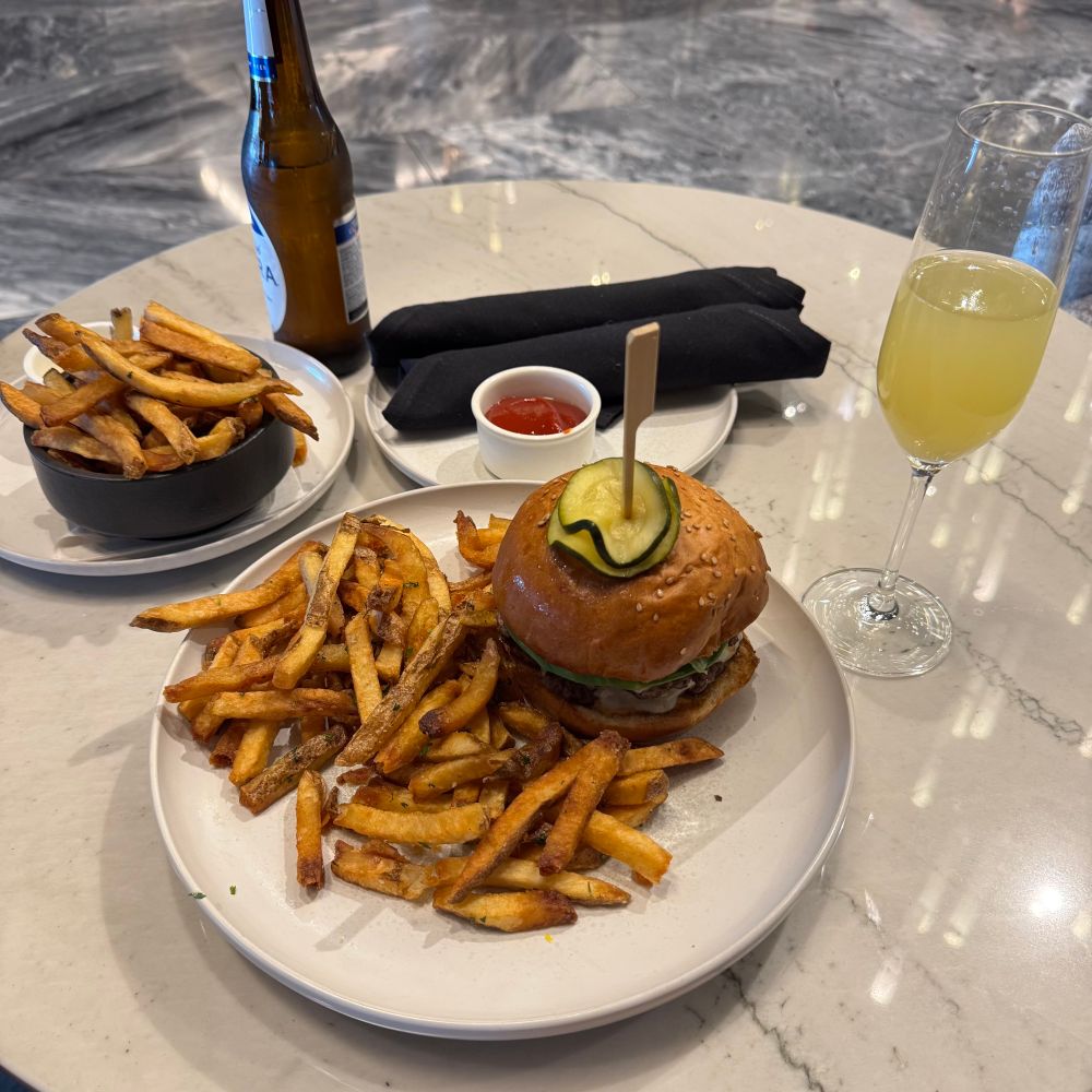 A plate of fries and a cheese burger with pickles poked on top. A Michelob Ultra and a mimosa on the table. 