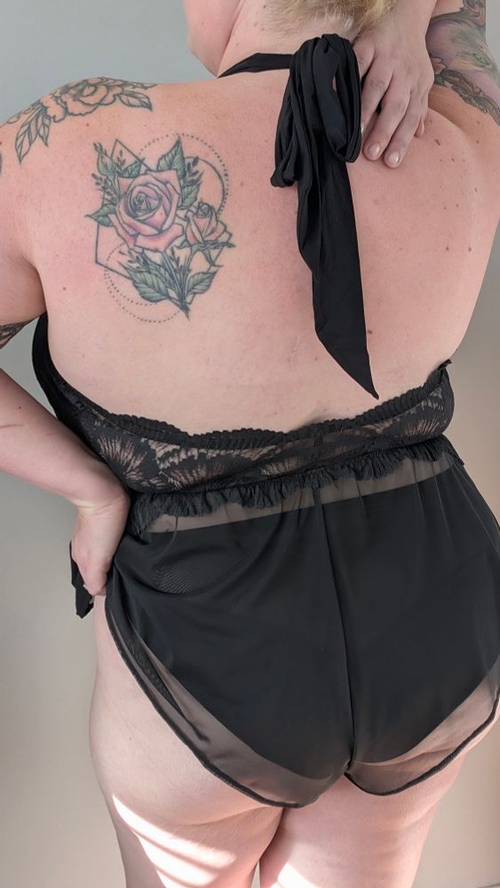 A photo from behind of a woman standing, wearing a black lingerie romper