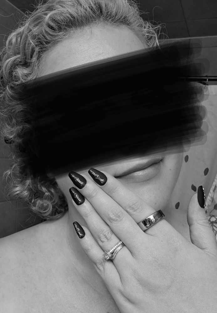 A black and white photo of a woman from shoulder up, with her hand up near her face, showing off long nails and silver-toned rings. Her face is censored.