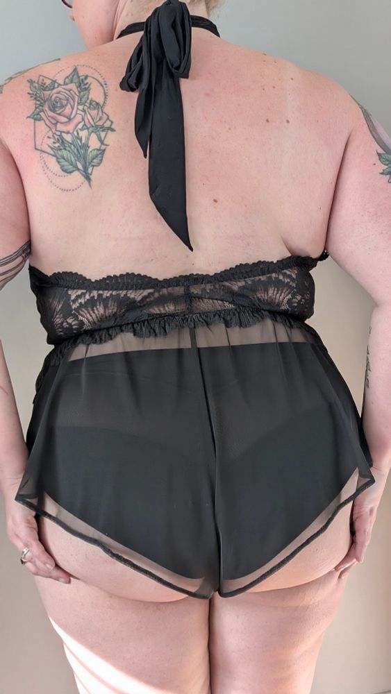 A photo taken from behind of a woman wearing a black lingerie romper and cupping her bottom with her hands on either side