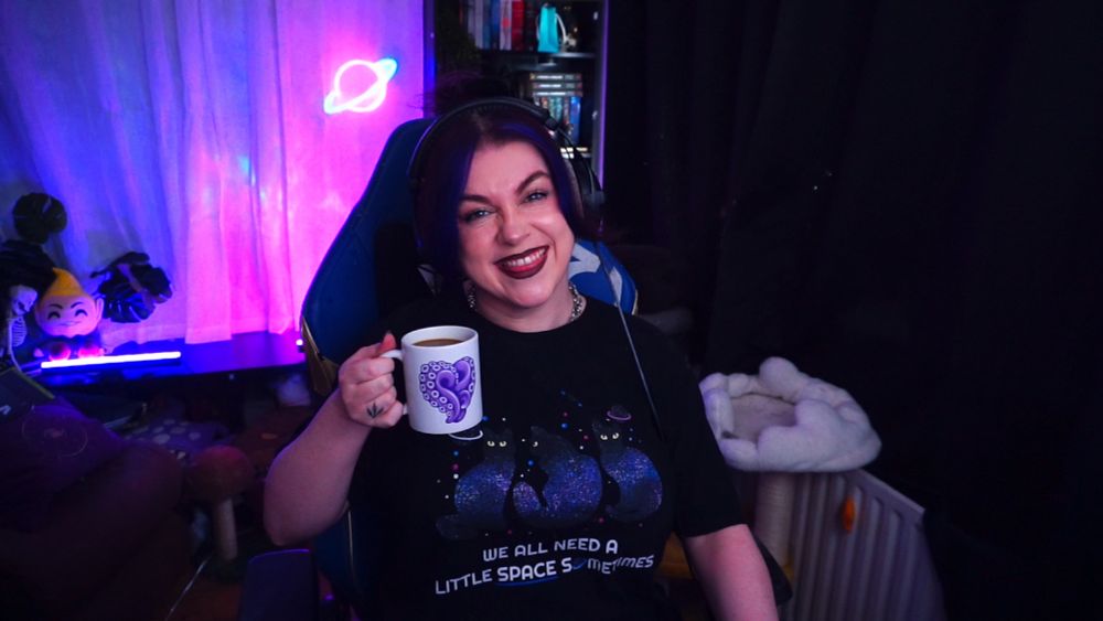 Psyche grins with her nose scrunched up. She's wearing a black t-shirt with three cat silhouettes on it, decorated with galaxy, star, and planet designs. She holds up a white mug with a purple heart formed from tentacles on it.