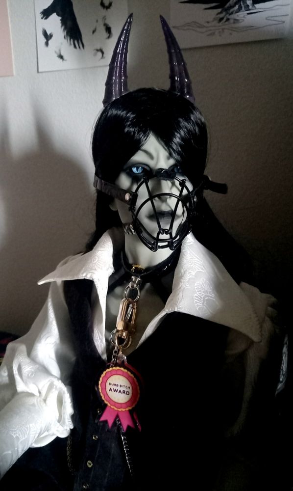 Ball jointed doll styled as a demon, sitting in dramatic lightning and wearing a muzzle, carrying a prize rosette that says "dumb bitch award)
