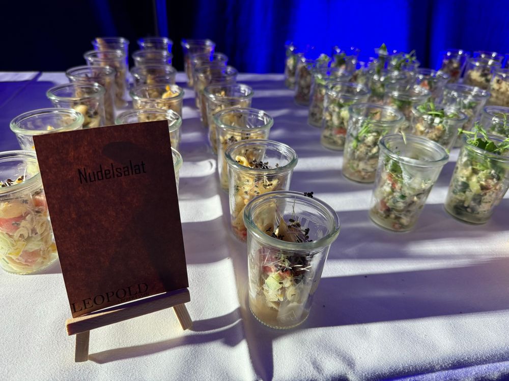 Das Bild zeigt kleine Gläser aufgereiht auf einem Tisch. In diesen Gläser befindet sich Nudelsalat oder auch normaler, kleingeschnittener Salat. Dies ist Fingerfood, bereitgestellt von einem Catering Unternehmen 