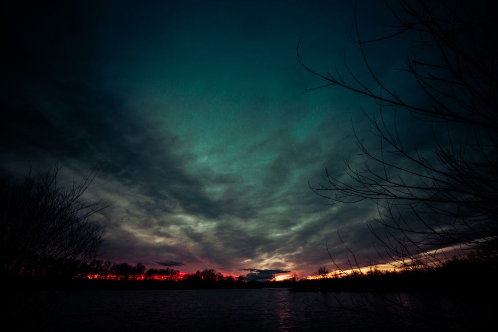 Wir sehen einen wolkenverhangenen Sonnenaufgang mit eine großen baumreihe rund um einen See am Morgen