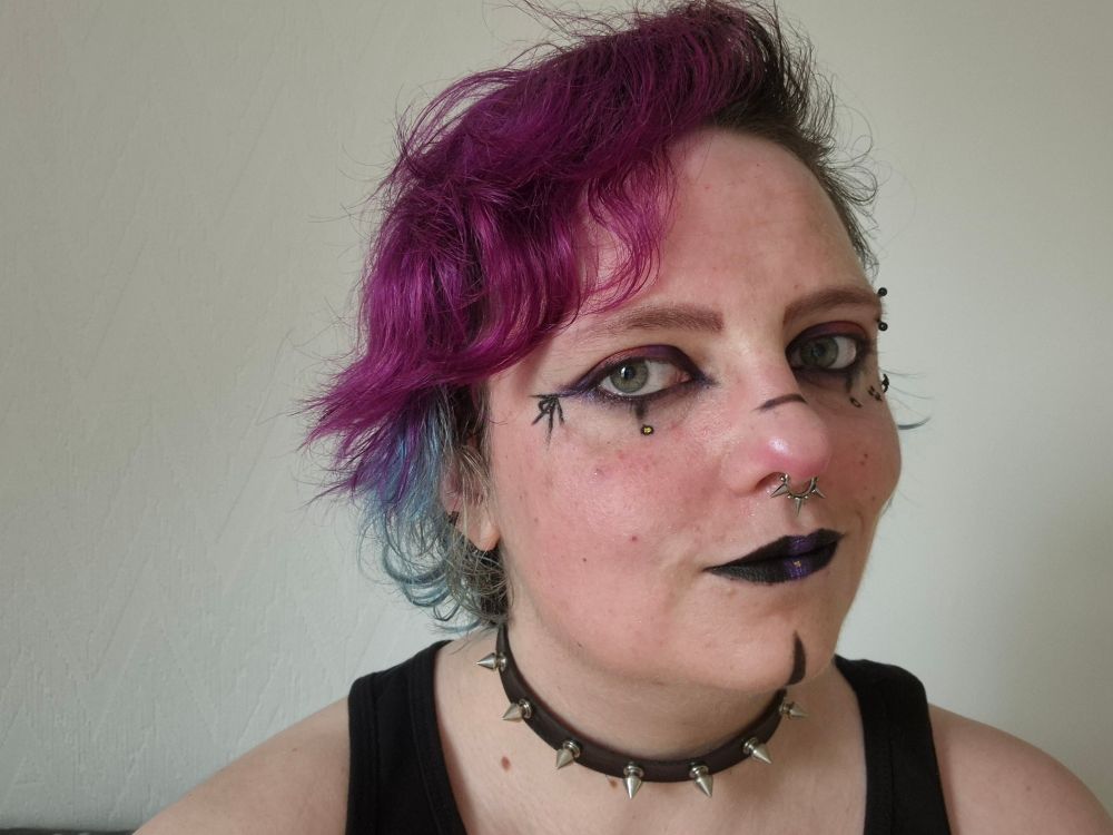 The hair is much longer now, vibrant purple on top, blue underneath, they have The Apocalypse Players monogram next to their eye and some Jesterish elements 