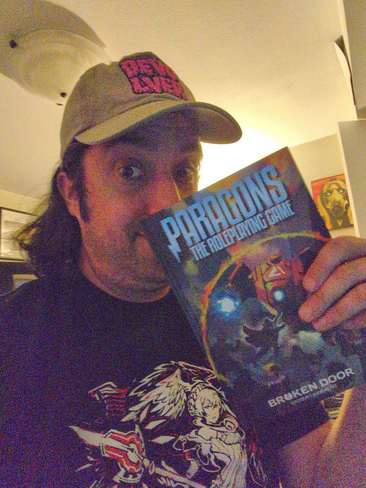 Lou holding a copy of Broken Door Entertainment's game, Paragons: The Roleplaying Game.