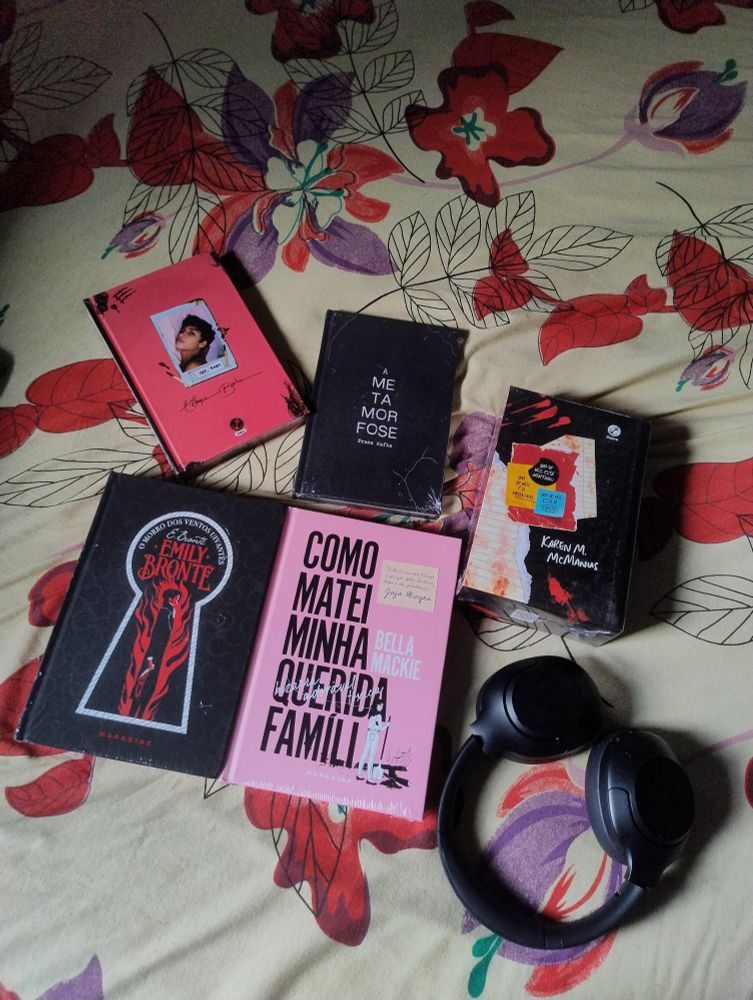 foto com um par de fones de ouvido Haylou S30; 1 box de livros da trilogia Um de Nós, da Karen M. McManus; livro Como Matei Minha Querida Família, de Bella Mackie; livro A Metamorfose, de Franz Kafka; livro Oxe, Baby, de Elayne Baeta: livro O Morro dos Ventos Uivantes, de Emily Brontë.