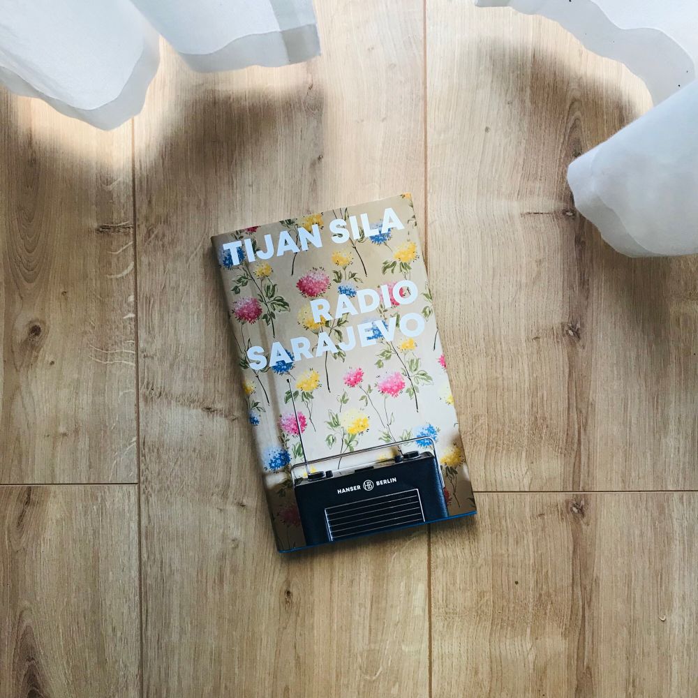 Auf dem Boden liegt das Buch „Radio Sarajevo“ von Tijan Sila. Auf dem Cover ist ein altes Radio zu sehen, dahinter eine geblümte Tapete.