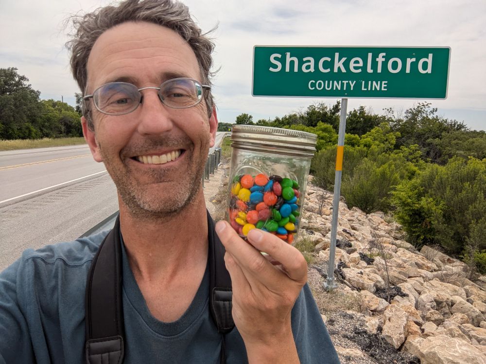 Photo of the author in front of a sign that says "Shackleford county line" holding a jar of M&Ms