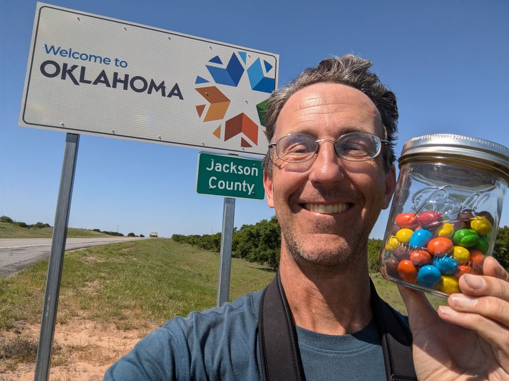 A photo of the author holding a jar of peanut M&Ms in front of a sign that says "Welcome to Oklahoma: Jackson County."