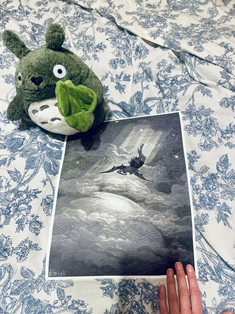 a photo of a black and white print of gustave doré’s engraving “fall of satan,” in which a winged figure falls dramatically downwards from celestial heights in a beam of divine light. the print is held in place by my hand and a small totoro stuffie