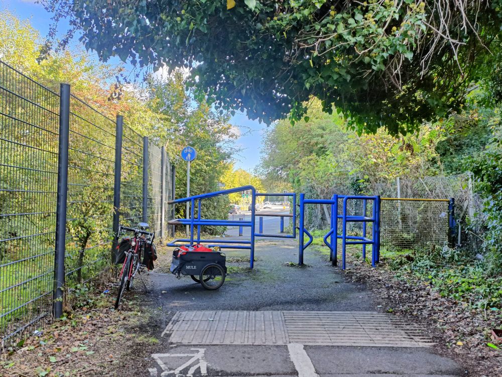 A shared cycle and pedestrian track with a white line separating the two sides. On the left is a metal fence, the right is lined with a wire mesh fence behind which is extensive vegetation. Leaf mulch and vegetation encroach on the path wodth from both sides. A cycle and trailer load of shopping are parked on the cycle side. Behind them is a blue tangle of metal resemblin a child's climbing frame and activity gym. There is a narrow gap between this and the fence.