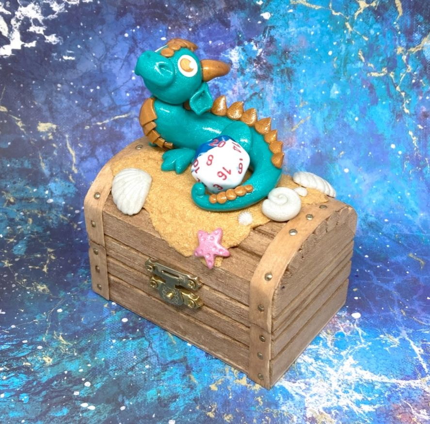 Photo of a polymer clay figure attached to a small wooden box.
The figure is that of a teal dragon, its tail curled up around a D20 die.
Clay sand and shells surround the dragon.