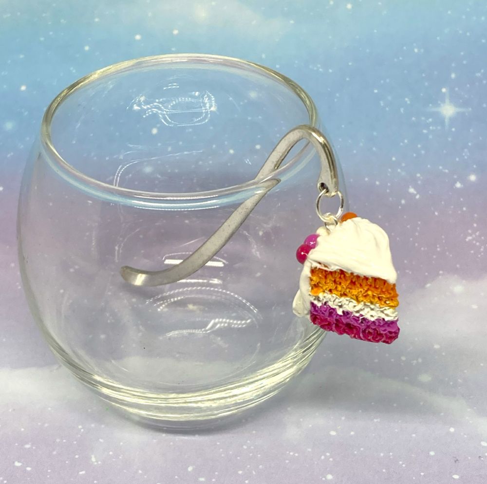 Photo of a polymer clay charm on a silver bookmark hook propped up inside a glass.
The charm is that of a slice of cake with the lesbian flag colors as the inside and white frosting.