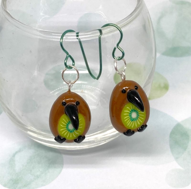 Photo of two polymer clay charms as earrings dangling off the rim of a glass.
The charms are that of kiwi birds with a slice of kiwi fruit in their bellies.