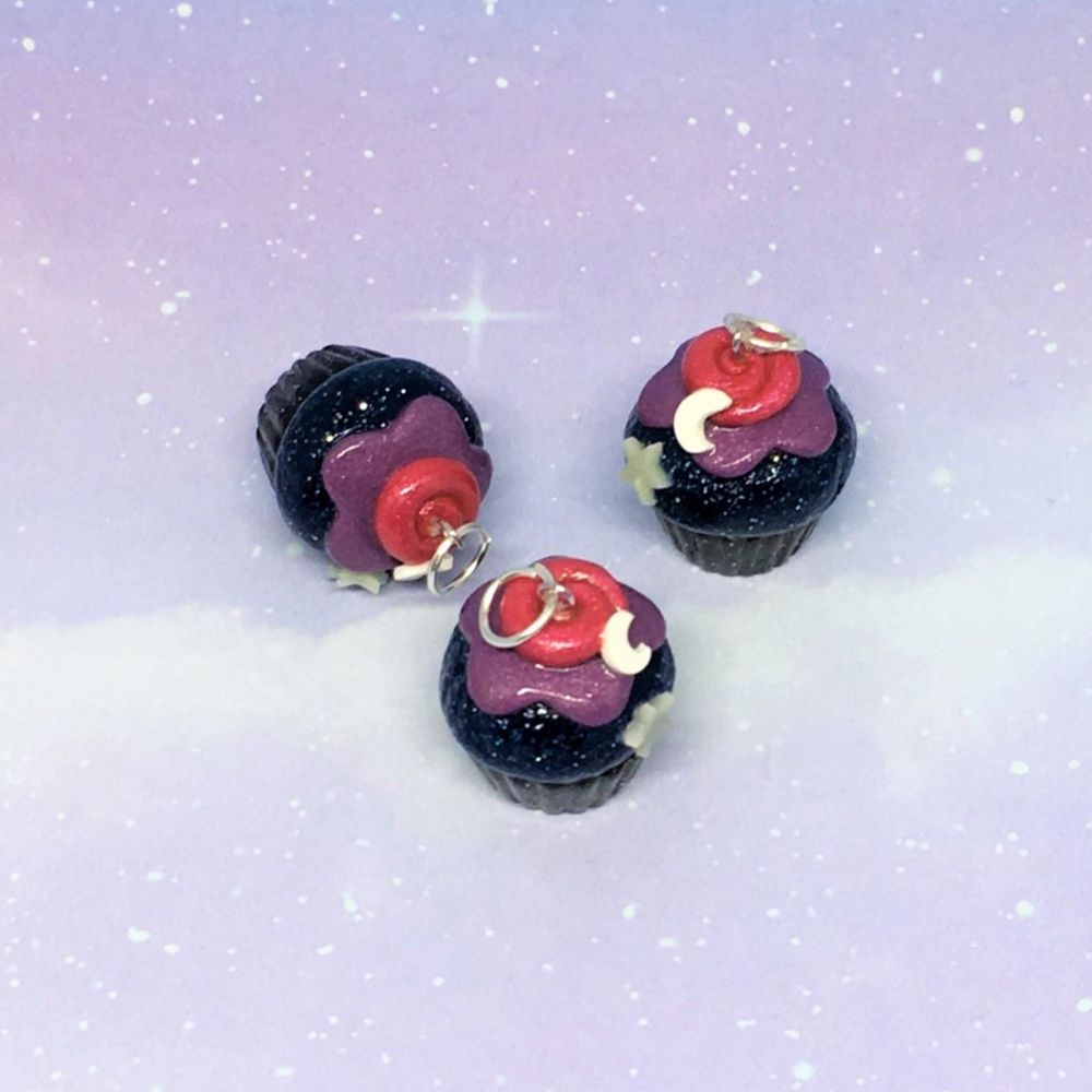 Three polymer clay cupcake charms.
The bottoms are sparkly black, the top is dark sparkly blue with sparkly purple frosting. A swirl of pink is on top with a crescent moon and glow int he dark star on each. Each is facing a different direction.