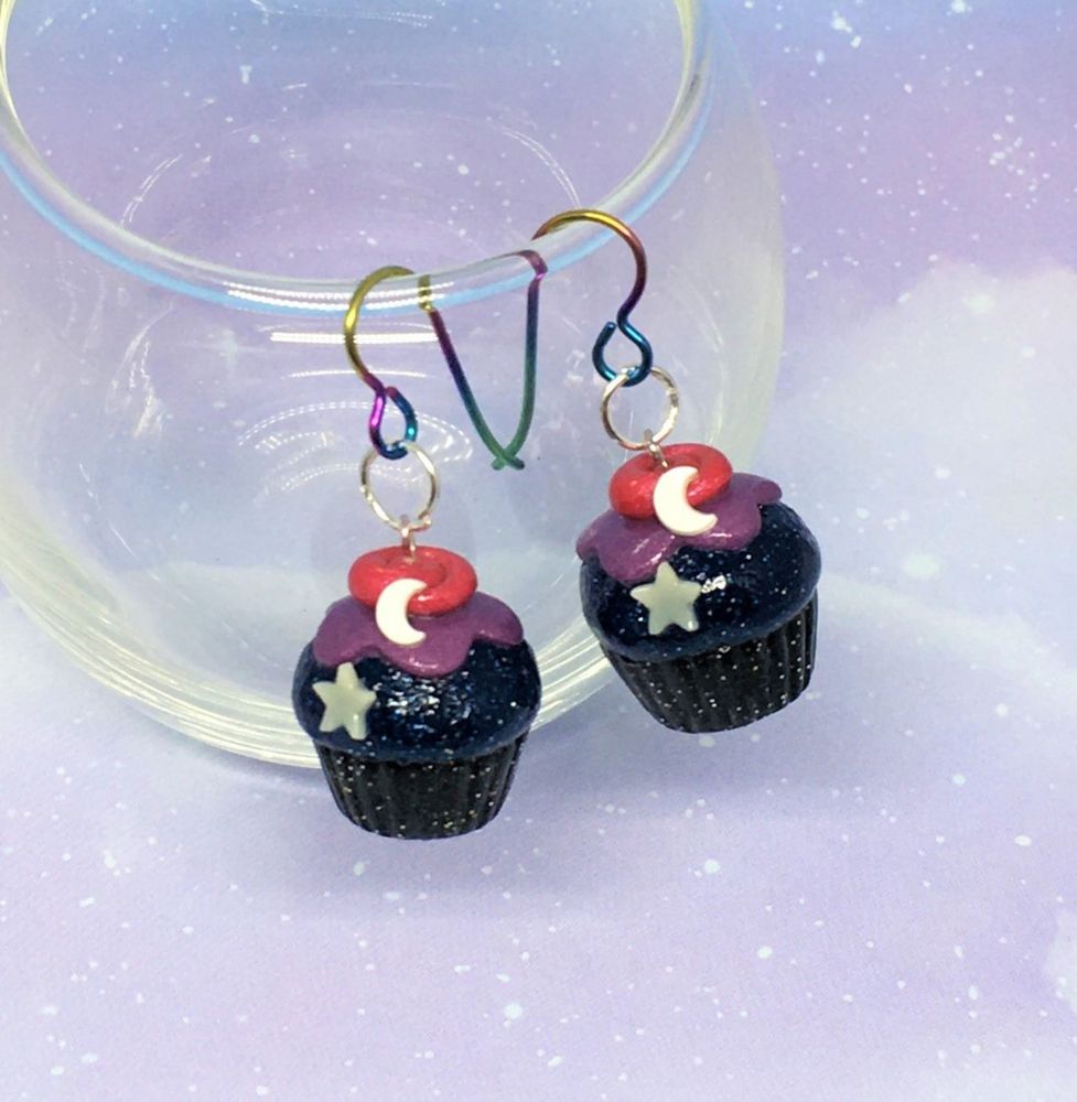 Two polymer clay cupcake charms on rainbow earring hooks dangling from the rim of a glass.
The bottoms are sparkly black, the top is dark sparkly blue with sparkly purple frosting. A swirl of pink is on top with a crescent moon and glow in the dark star on each. 
