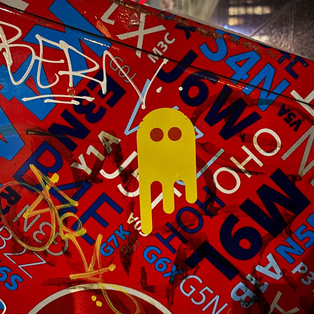A Melting Ghost sticker, yellow with red eyes, on a red Canada Post mail-collection box. The pattern on the box is Canadian postal codes set in black, white, and blue, on diagonals. The box’s surface is also covered in graffiti. 